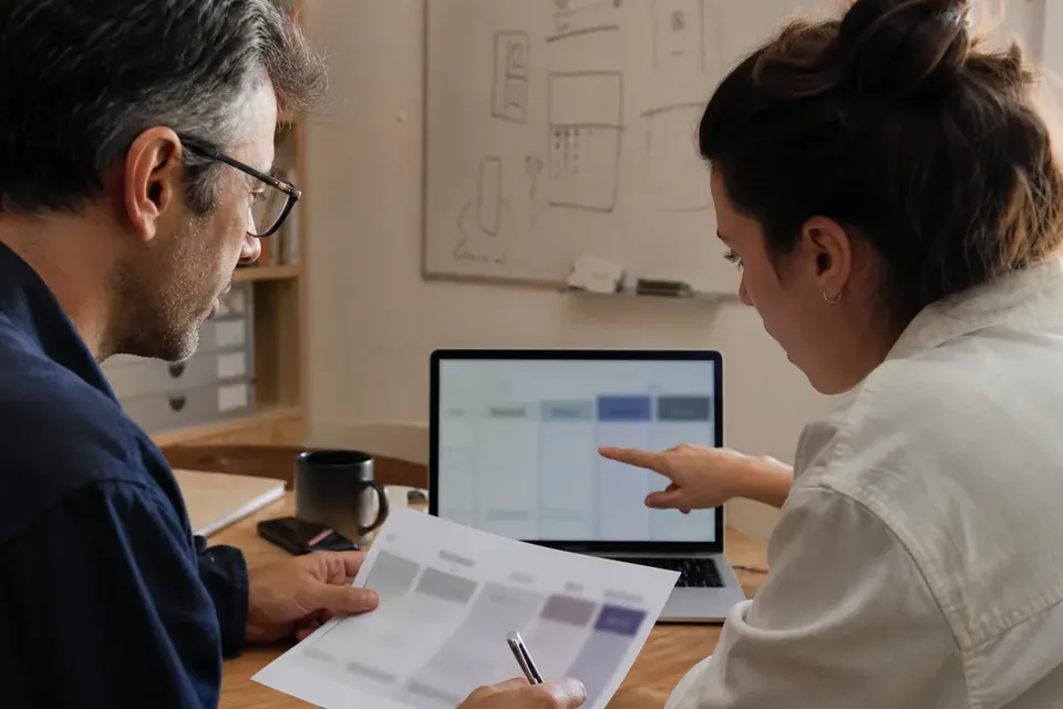 Two founders comparing a printed fee schedule with a laptop at a shared desk