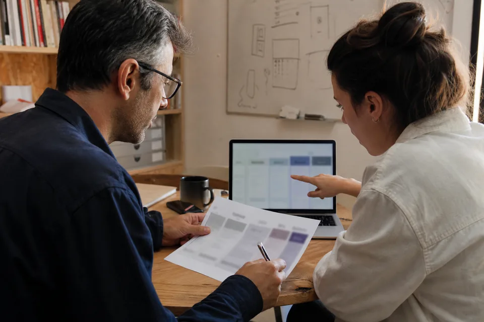 Two startup co-founders comparing business banking plan costs on a printed sheet and a laptop